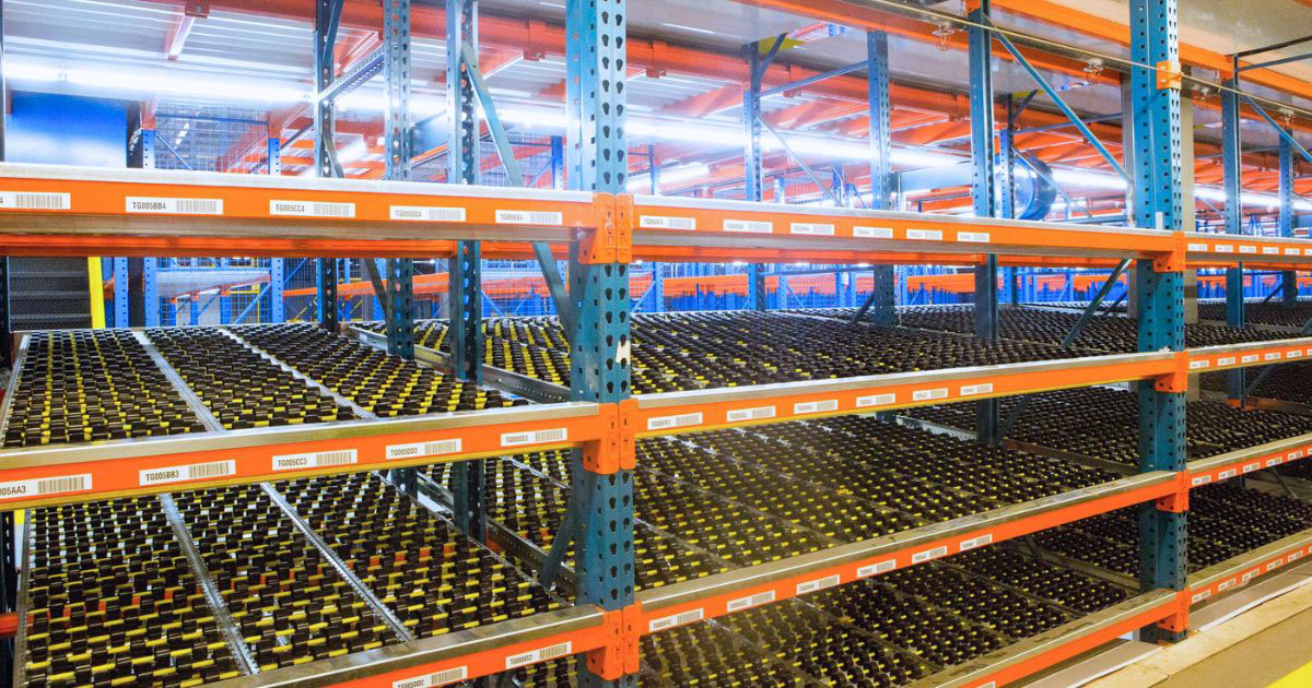 An elevated view of a carton flow racking system featuring blue vertical uprights and orange horizontal beams. The levels are equipped with gravity-fed roller tracks designed for small-item picking.