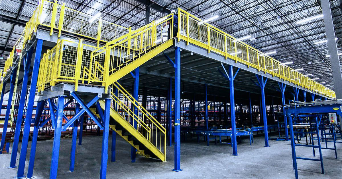 A wide-perspective shot of a heavy-duty industrial steel mezzanine system. The structure features blue support columns and beams topped with a gray decking platform, accessible via a bright yellow staircase.