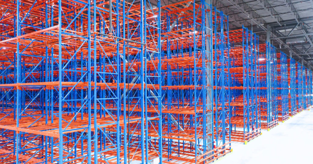 A wide shot of a vast warehouse filled with rows of blue and orange industrial selective pallet racking systems under bright overhead LED lighting.