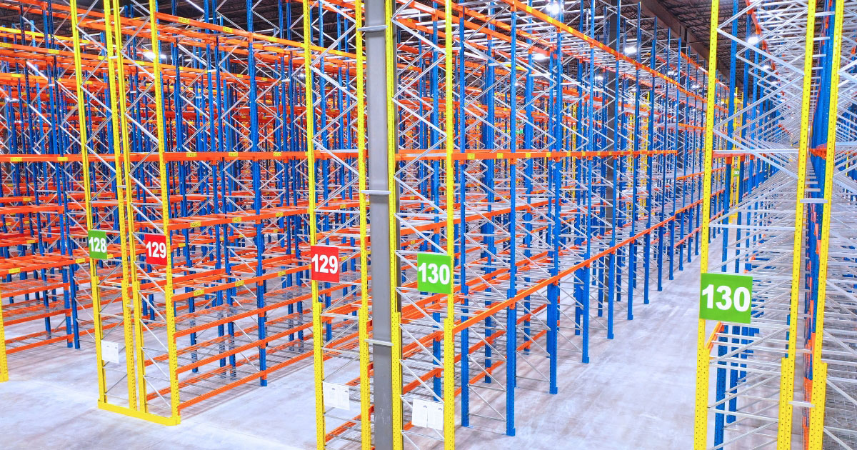 A high-perspective view of a massive, empty selective racking system featuring blue and yellow frames, orange beams, and numbered aisle signs stretching across a polished warehouse floor.